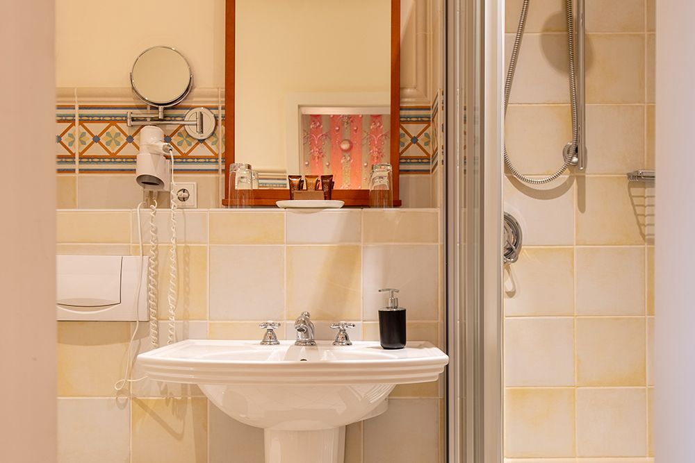 Badezimmer mit Waschbecken, Seifenspender, Föhn und Spiegel. Dusche mit Schiebetür rechts. Geflieste Wände und Boden.