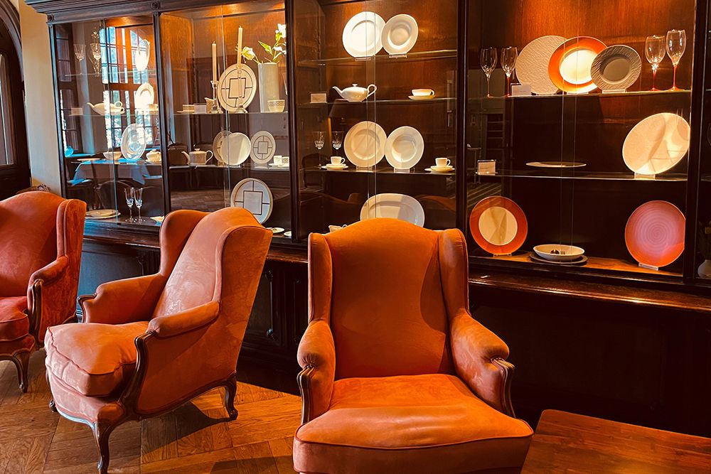 Zwei orangefarbene Sessel stehen gemütlich in einem kleinen Esszimmer gegenüber einer Glasvitrine, in der verschiedene Essgeschirre ausgestellt sind – Teller, Tassen und Untertassen. Der Holzboden unter den Füßen ergänzt die gedämpfte Beleuchtung und schafft einen perfekten Ort, um Ihren Morgenkaffee zu genießen.