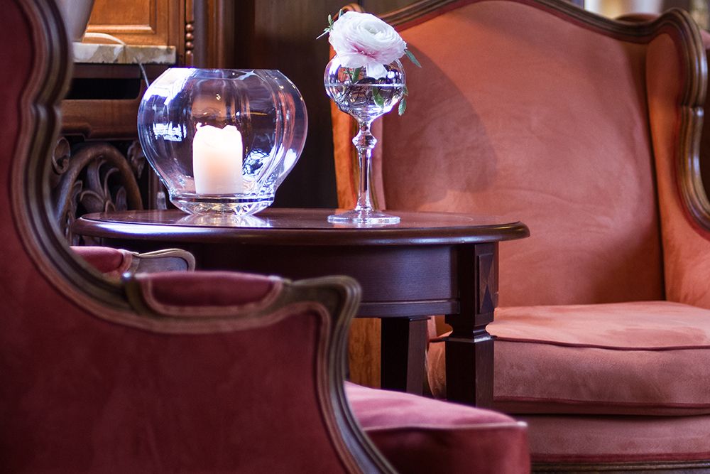 Zwei rote Samtstühle flankieren einen Holztisch mit Parkettboden darunter. Auf dem Tisch steht eine brennende Kerze in einem Glashalter und eine einzelne weiße Rose in einer Vase, was den Charme eines intimen Kaminzimmers hervorruft.