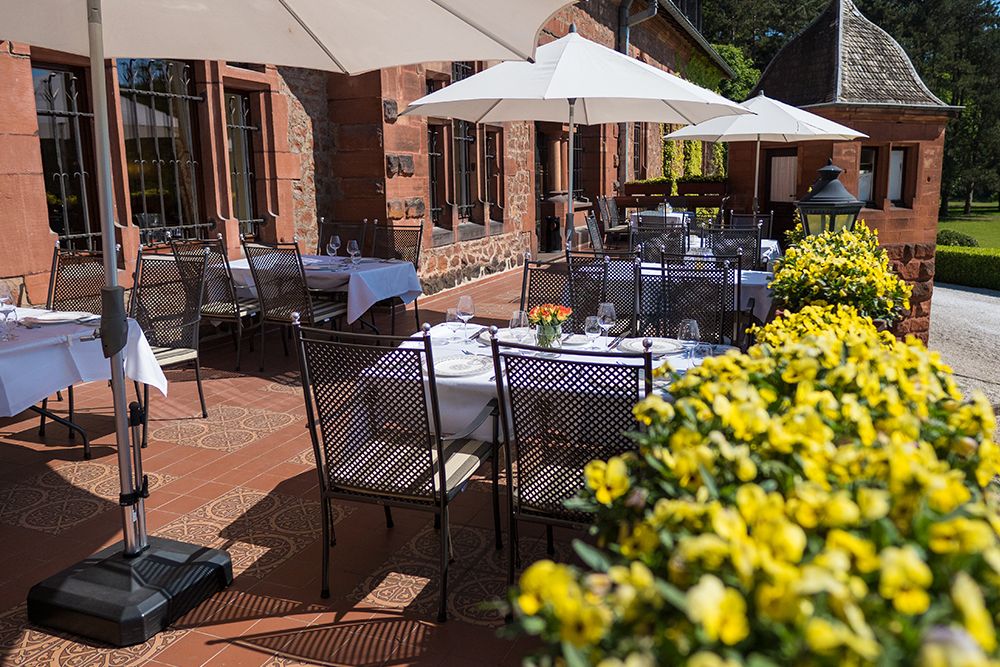 Die bezaubernde Außenterrasse, die sogenannte Schloss-Terrasse, bietet Tische mit weißen Tischdecken und ist von leuchtend gelben Blumen umgeben. Im Schatten eleganter Sonnenschirme liegt sie anmutig neben einem Backsteingebäude und bietet eine ruhige Parkatmosphäre in der Nähe der malerischen Saar.