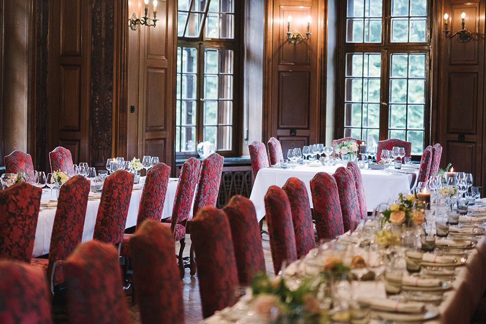 Elegantes Esszimmer mit zwei langen Tischen, die für eine formelle Veranstaltung gedeckt sind. Die Tische sind mit weißen Tischdecken, Glaswaren und Blumenschmuck ausgestattet. Die Tische sind mit roten Polsterstühlen gesäumt, was es perfekt für Hochzeiten macht. Große Fenster im Hintergrund vervollständigen das anspruchsvolle Ambiente.