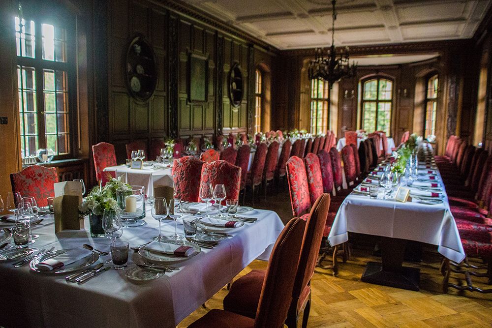 Ein formelles Esszimmer, perfekt für Hochzeiten, mit zwei langen Tischen mit weißen Tischdecken und roten Stühlen. Die Gedecke sind elegant arrangiert, umgeben von reicher Holzvertäfelung und großen Fenstern, die den Raum mit natürlichem Licht durchfluten.