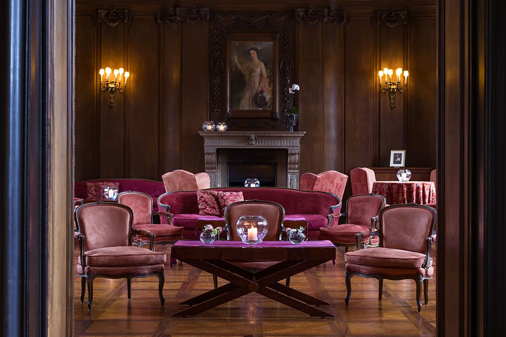 Elegantes Kaminzimmer mit Holzwänden, einem Kamin und einem Gemälde darüber. Samtstühle und Sofas sind um dunkle Holztische angeordnet, perfekt zum Nippen an Single Malt Whiskey. Sanftes Licht von Wandleuchten schafft eine warme Atmosphäre.