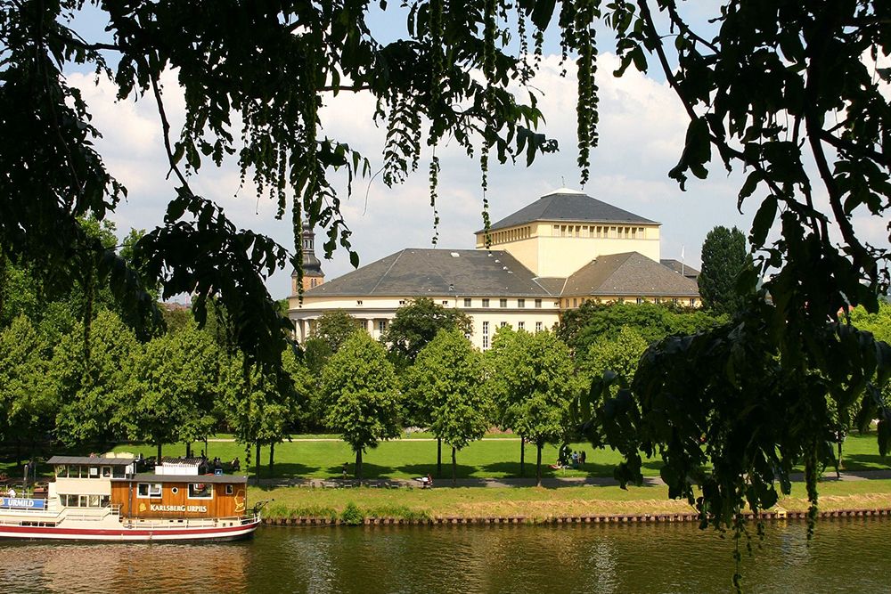 A boat passes by a river, a large building can be seen in the background, framed by trees and greenery under a clear sky.