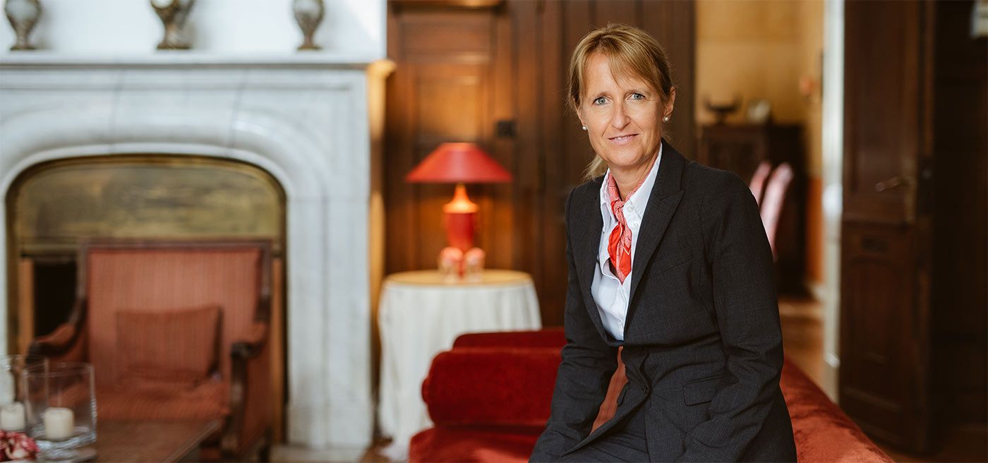 Eine Person im Anzug mit rotem Schal sitzt auf einer Couch im gemütlichen Ambiente von Schloss Saareck, wo das warme Leuchten des Kamins und einer Vintage-Lampe eine einladende Atmosphäre schaffen.