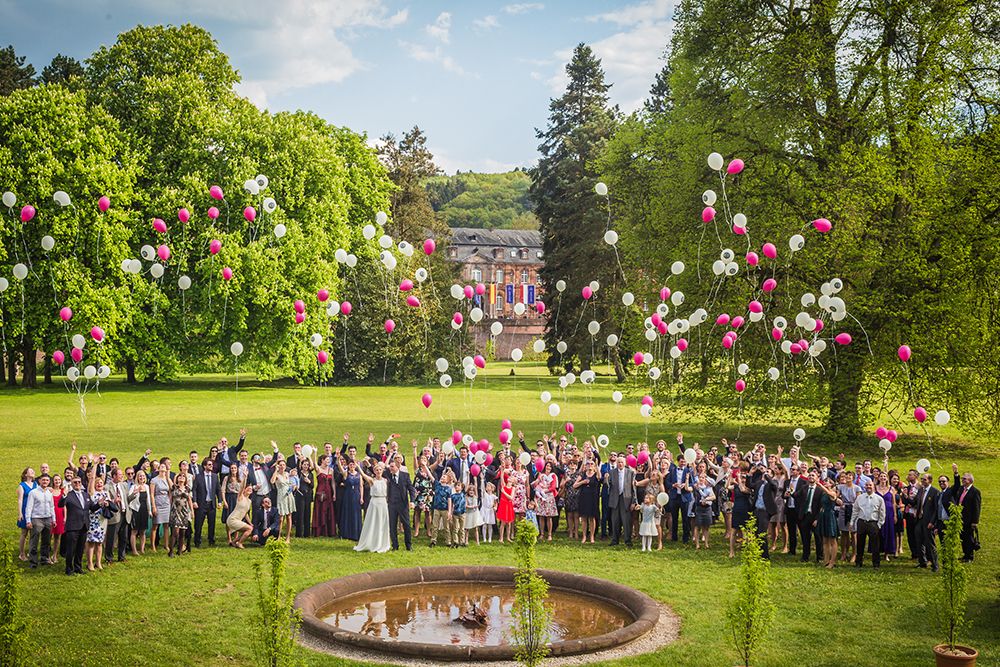 Eine große Gruppe von Menschen steht im Freien und lässt rote und weiße Luftballons in den Himmel steigen. Ein Brunnen und ein majestätisches Schloss bilden eine märchenhafte Kulisse, umgeben von grünen Bäumen und Gras.