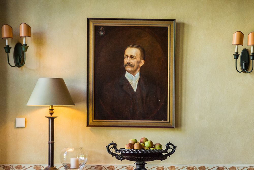 An der Wand hängt ein gerahmtes Porträt eines Mannes mit Schnurrbart. Darunter schmücken eine Lampe, eine Glasschale und eine Obstschale mit Äpfeln den Tisch und erinnern an ein elegantes Frühstücksbuffet. Zwei Wandlampen sorgen auf beiden Seiten für Licht.