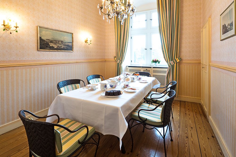 Im eleganten Speisesaal mit Blick auf den Saareckpark ist unter einem kunstvollen Kronleuchter ein langer Tisch für den Tee gedeckt. Gestreifte grüne Vorhänge rahmen den Blick auf die Saarseite ein, während hellrosa Wände mit dekorativen Paneelen und gerahmten Kunstwerken diesem exquisiten Raum Charme verleihen.