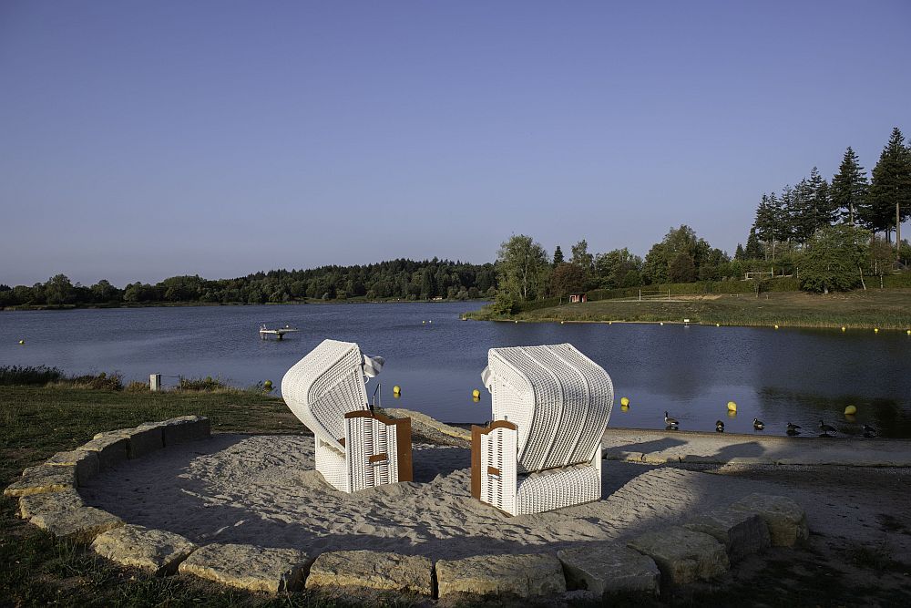 Two white beach chairs on a sandy beach overlooking a calm lake with trees in the background under a clear sky.