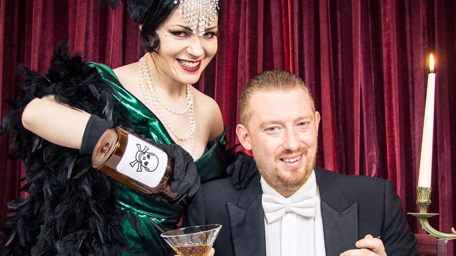 Vor einem roten Vorhang gießt eine Frau im Vintage-Outfit, die Teil des kulinarischen Programms auf Schloss Saareck ist, Flüssigkeit aus einer Totenkopfflasche in das Glas eines Mannes. Beide sind formell gekleidet und lächeln schelmisch.