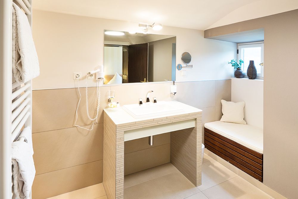 Modernes Badezimmer mit Waschbecken, großem Spiegel, wandmontiertem Haartrockner, Handtüchern und einer gepolsterten Bank unter einem kleinen Fenster.