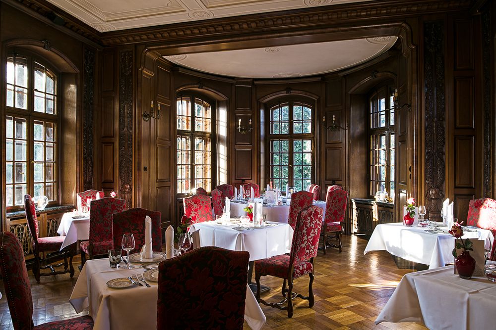 Elegantes Esszimmer mit Holzvertäfelung, großen Bogenfenstern und roten Polsterstühlen, perfekt für stilvolle Hochzeiten. Die Tische sind mit weißen Tischdecken, Gläsern und gefalteten Servietten gedeckt und schaffen ein anspruchsvolles Ambiente.