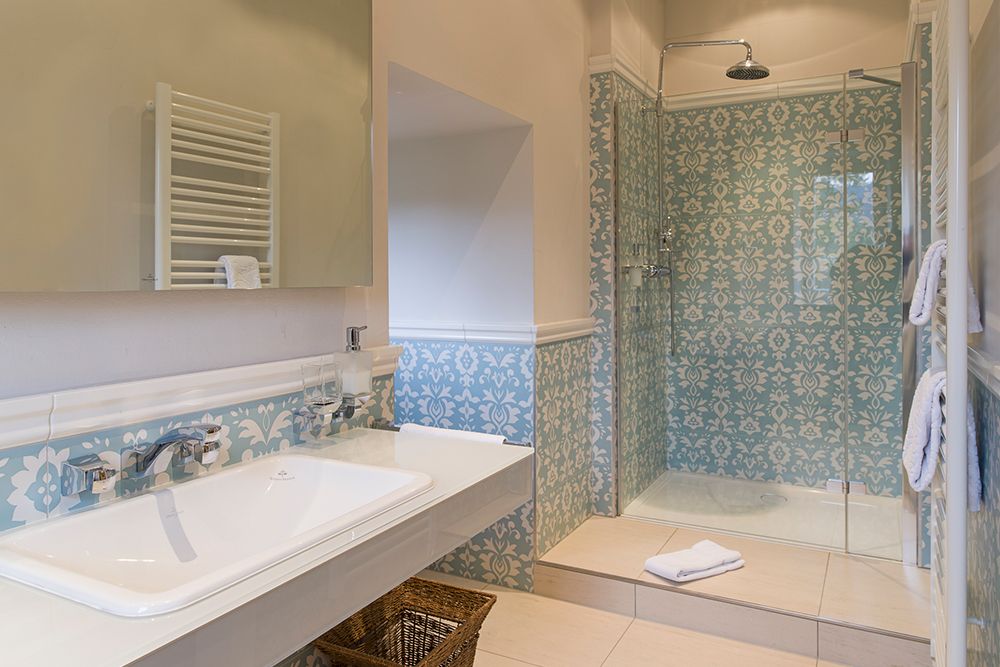 Badezimmer mit großem Waschbecken, gemusterter gefliester Dusche, Glastür, wandmontiertem Handtuchhalter und Aufbewahrungskorb aus Weidengeflecht.