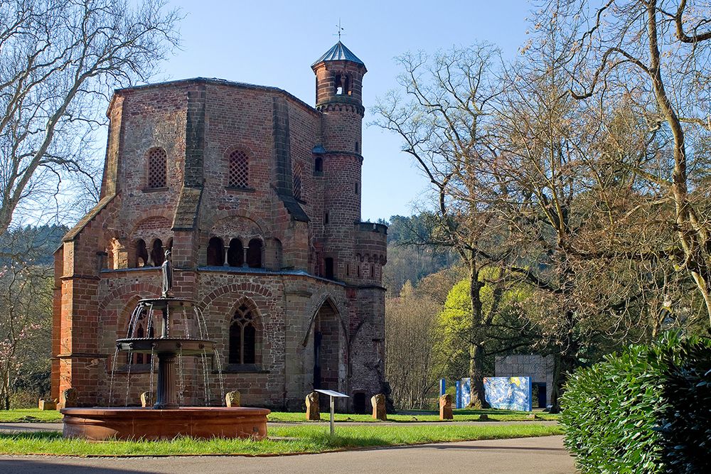 Known for its round arches and square bell tower, this red brick church is nestled among trees and greenery under a clear blue sky. It is ideal for day trips and conveniently located near hotels. A round fountain adorns its front.
