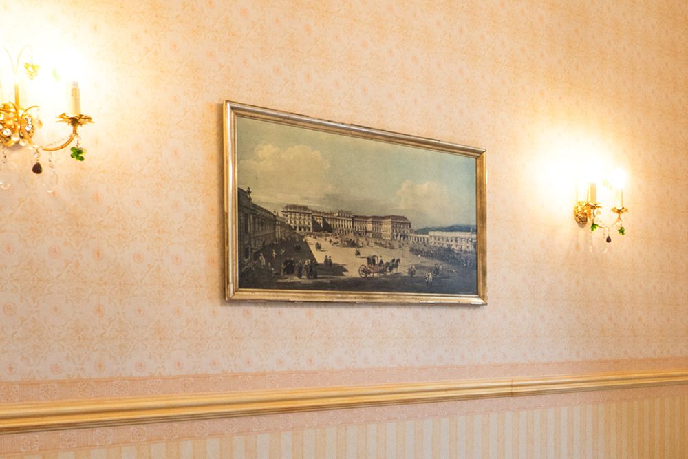 An einer Wand mit floraler Tapete, die an das Gelbe Zimmer erinnert, hängt ein gerahmtes Gemälde mit einer historischen Gebäudeszene. Zwei verzierte Wandleuchter mit kleinen grünen Verzierungen flankieren das Gemälde.