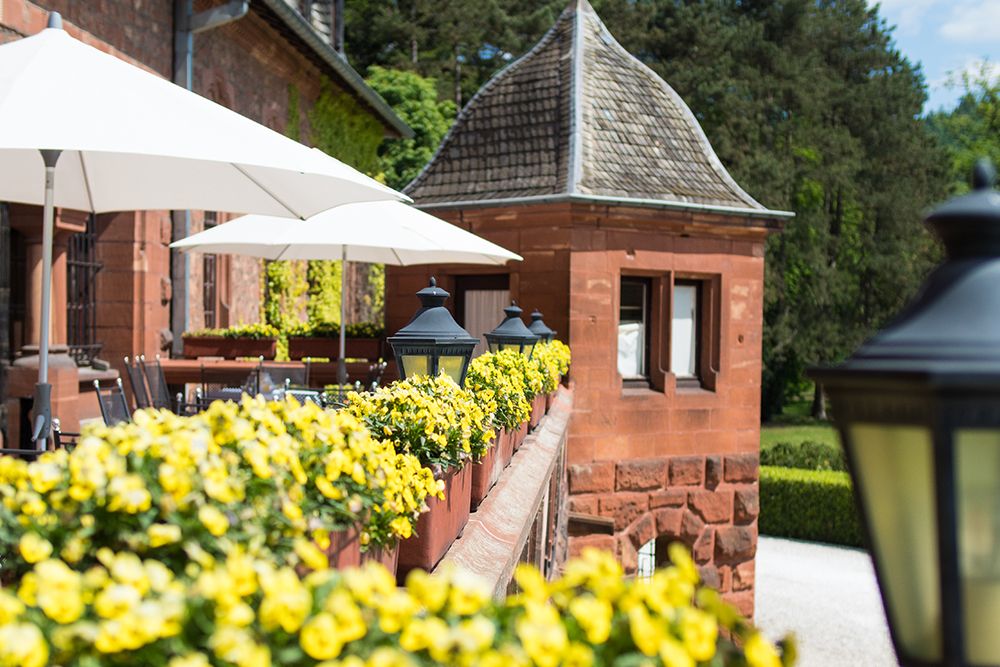 Die Schloss-Terrasse ist eine bezaubernde Terrasse mit weißen Sonnenschirmen und gelben Blumen entlang eines Backsteingeländers. Neben diesem historischen Backsteingebäude mit seinem konischen Dach wird die ruhige Umgebung durch die üppigen Bäume des umliegenden Saarparks wunderbar ergänzt.