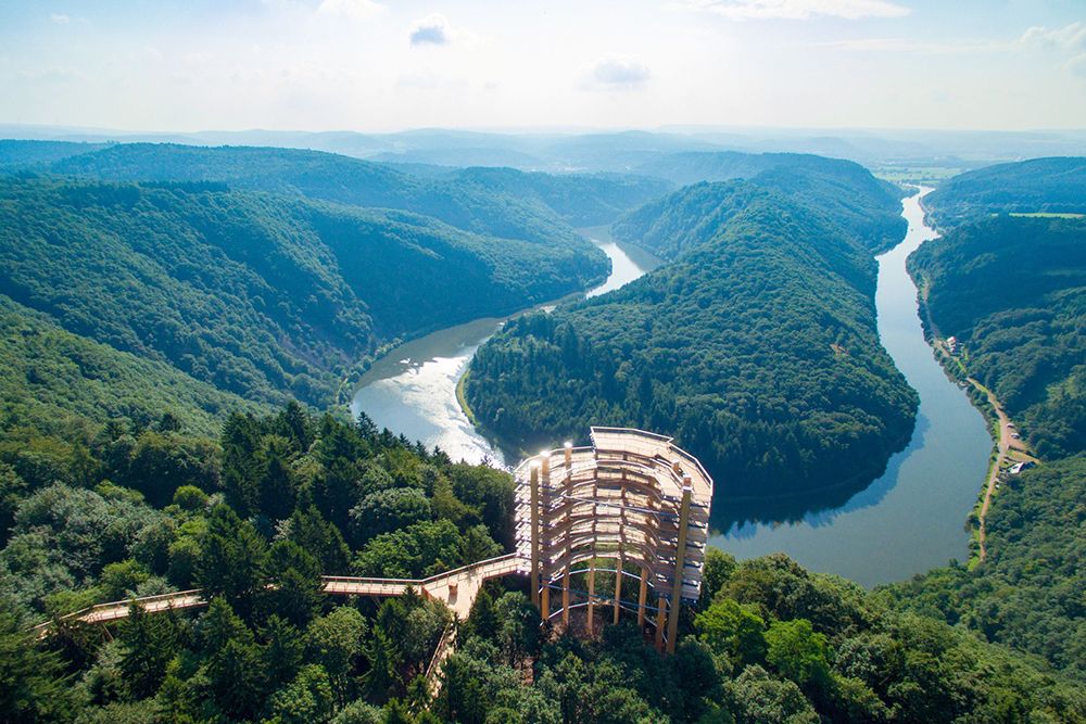 Wooden lookout tower in a lush, green forest overlooking a winding river and distant hills under a clear sky.