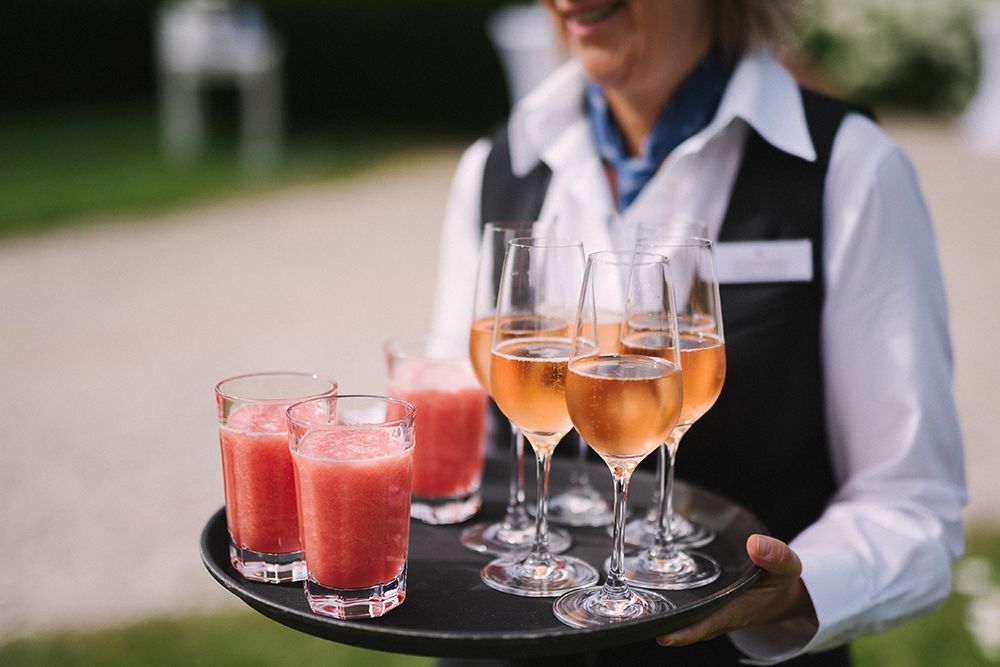 Ein Kellner im Schloss Saareck hält draußen ein Tablett mit vier Gläsern rosa Saft und vier Champagnergläsern mit einem rosa Getränk. Auf diese einmalig schöne Weise fühlt es sich wirklich so an, als wäre das Leben ein Fest.
