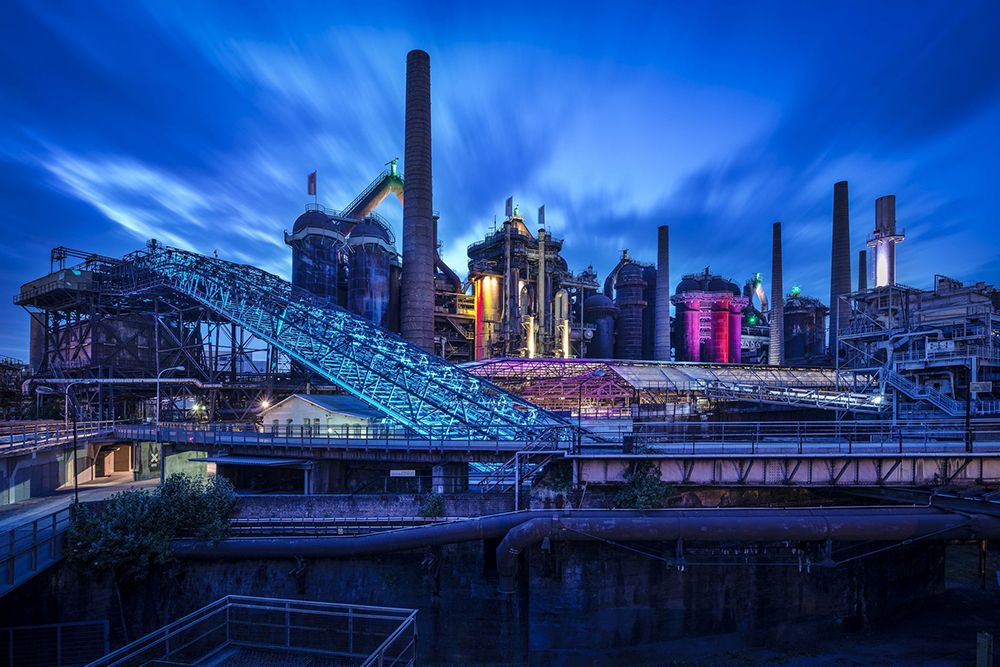 The Saarland industrial complex features illuminated structures, tall chimneys and a conveyor belt against a cloudy blue sky at dusk. This impressive scene is one of the region's most fascinating sights.