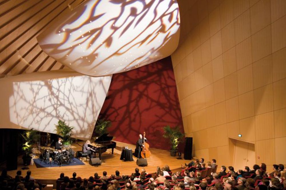 Ein Jazztrio verzaubert das Publikum bei den Kammermusiktagen. Auf der Bühne treten Klavier, Kontrabass und Schlagzeug auf. Künstlerische Lichteffekte werfen Schatten an die Decke des Kulturkonzertsaals, während Musik die Luft erfüllt.