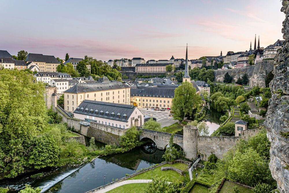 Die Luftaufnahme der Stadt Luxemburg zeigt ihre historischen Gebäude, eine Steinbrücke über den Fluss und üppiges Grün unter einem klaren Himmel – ein perfektes Ziel für Stadttouren-Enthusiasten.