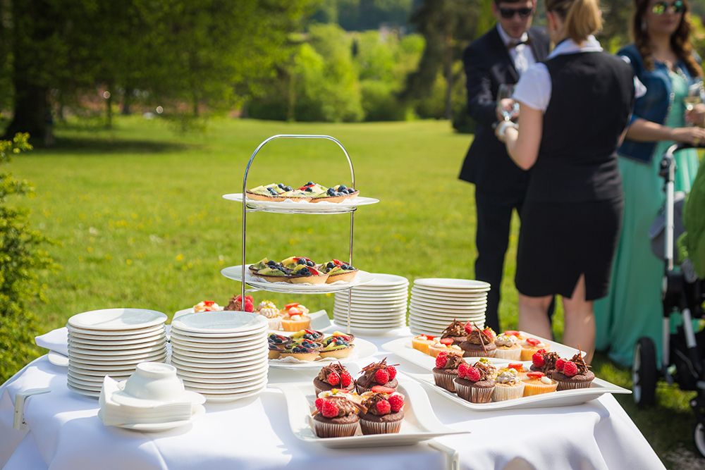Buffet table for an outdoor event with dessert plates, cupcakes and stacked plates. In the background, people chatting in a garden setting.