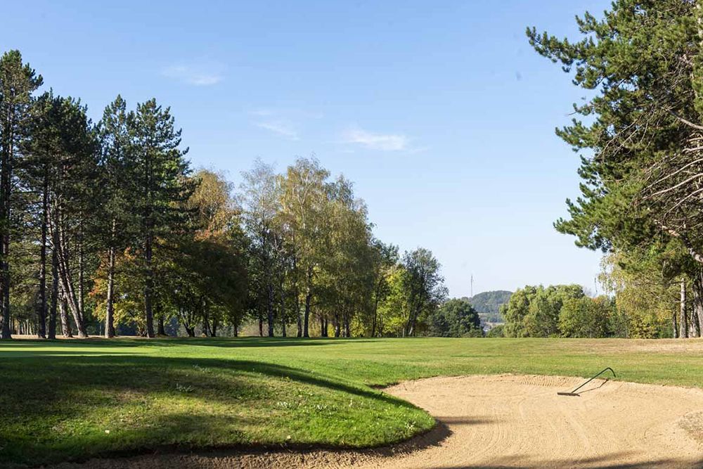 Ein Golfplatz mit einem Sandbunker im Vordergrund, umgeben von grünem Gras und Bäumen unter einem klaren blauen Himmel, ist die perfekte Umgebung, um Ihr Handicap zu verbessern.