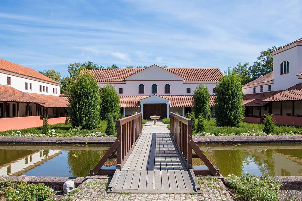 A wooden bridge leads to a Roman-style villa, one of Saarland's magical destinations, surrounded by a well-kept garden and a reflecting pond under a blue sky.