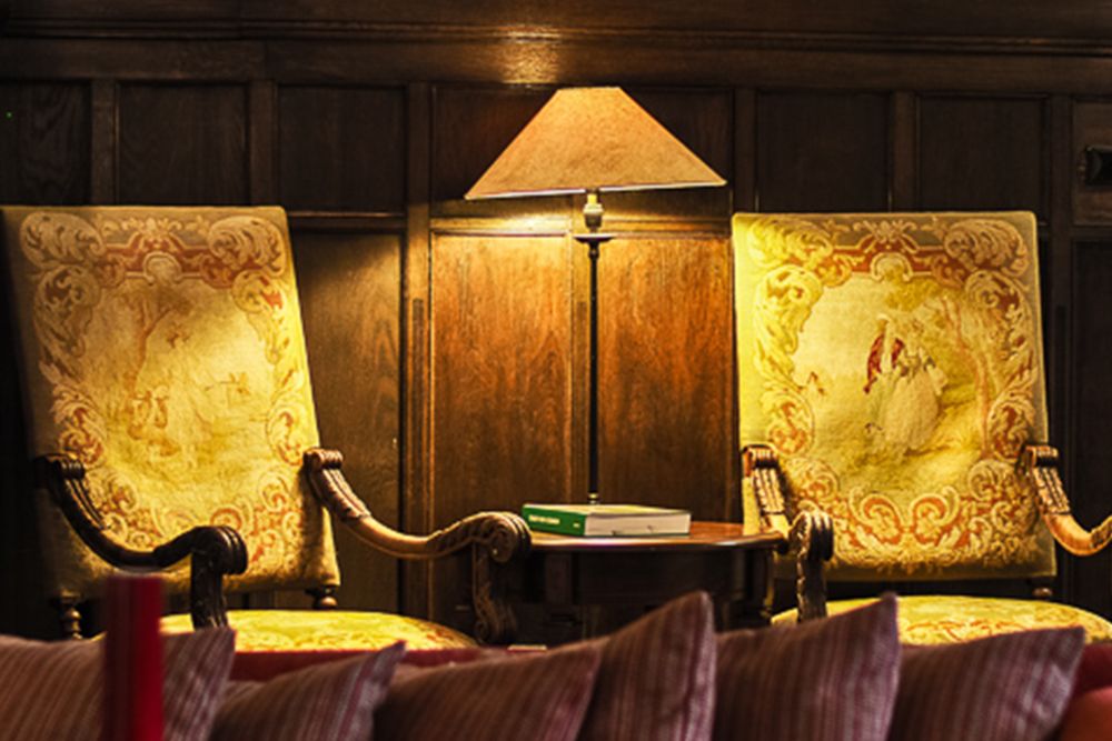 Zwei reich verzierte Polsterstühle mit komplizierten Mustern flankieren einen Holztisch, der sich perfekt für einen Aperitif mit einem Buch unter einer brennenden Lampe eignet, vor einer dunklen holzgetäfelten Wand in der gemütlichen Hotellobby.