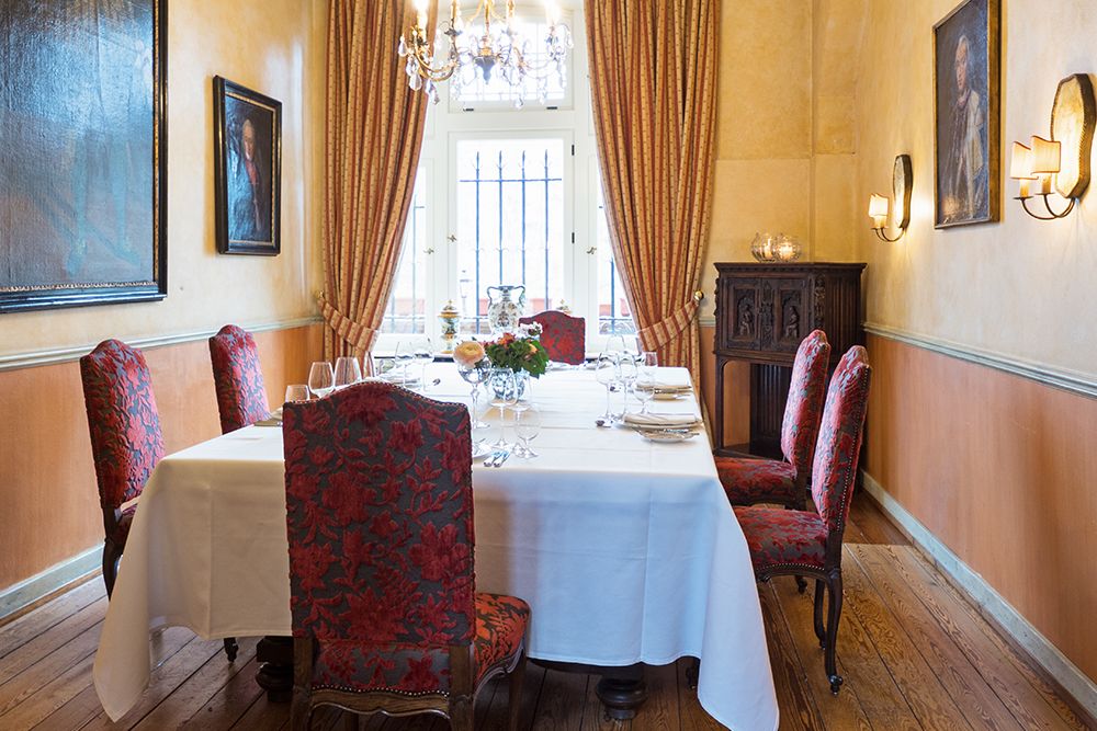 Elegantes Esszimmer in Straßburg mit einem für sechs Personen gedeckten Tisch. Rot gepolsterte Stühle, weiße Tischdecken und Holzboden schaffen eine stimmungsvolle Atmosphäre. Kronleuchter und Gemälde schmücken die Wände, während ein großes Fenster mit Vorhängen den Charme verstärkt, perfekt für jede Feier.