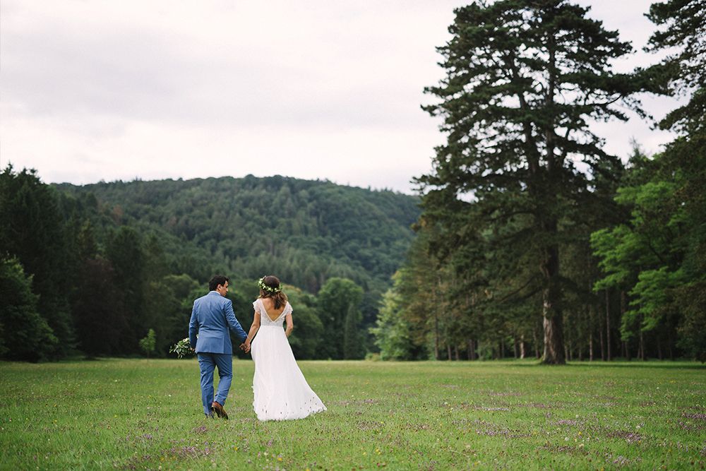 A couple, the groom in a blue suit and the bride in a white dress, hold hands while walking across a meadow with trees and hills in the background.