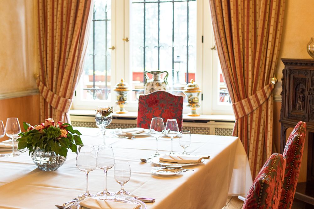 Elegantes Esszimmer mit gedecktem Tisch, weißer Tischdecke, ordentlich angeordneten Weingläsern, Tellern und einem Blumengesteck. Verzierte Vorhänge rahmen das große Fenster ein und schaffen eine stimmungsvolle Atmosphäre, die perfekt für jeden Raum ist, der für Feiern gedacht ist.