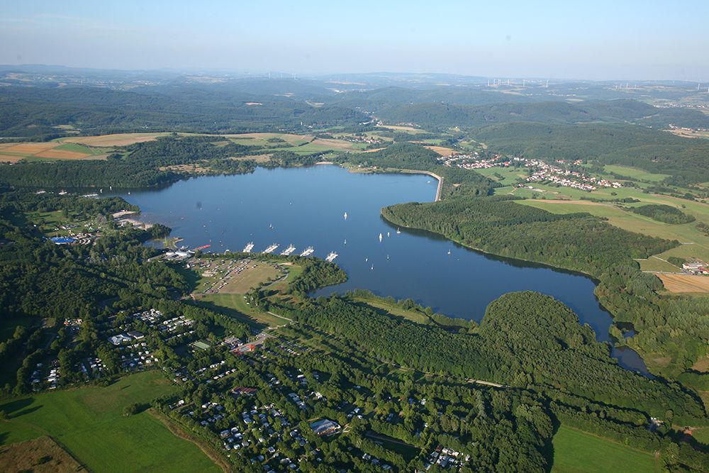 Aerial view of a huge lake surrounded by woods, fields and scattered buildings, boats can be seen on the water.