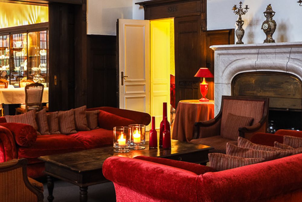 Gemütlicher Raum mit roten Sofas um einen Holztisch, brennenden Kerzen und einem Kamin erinnert an eine einladende Hotellobby. Eine offene Tür führt zu einem Essbereich, der sich perfekt für einen Aperitif eignet.