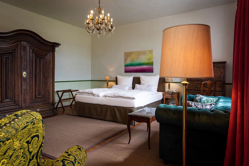 Gemütliches Hotelzimmer mit Kronleuchter, Doppelbett, Vintage-Möbeln, großer Lampe, Gemälde und buntem Sessel.