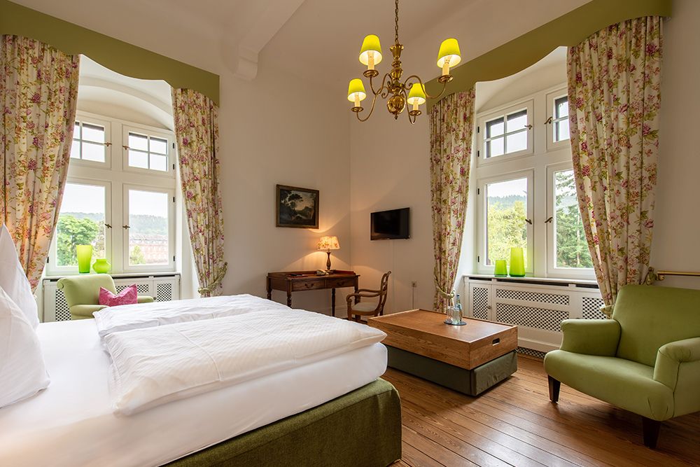 Helles Hotelzimmer mit Bett, Schreibtisch, Sessel und Fenstern mit geblümten Vorhängen. Kronleuchter und an der Wand montierter Fernseher sind sichtbar.
