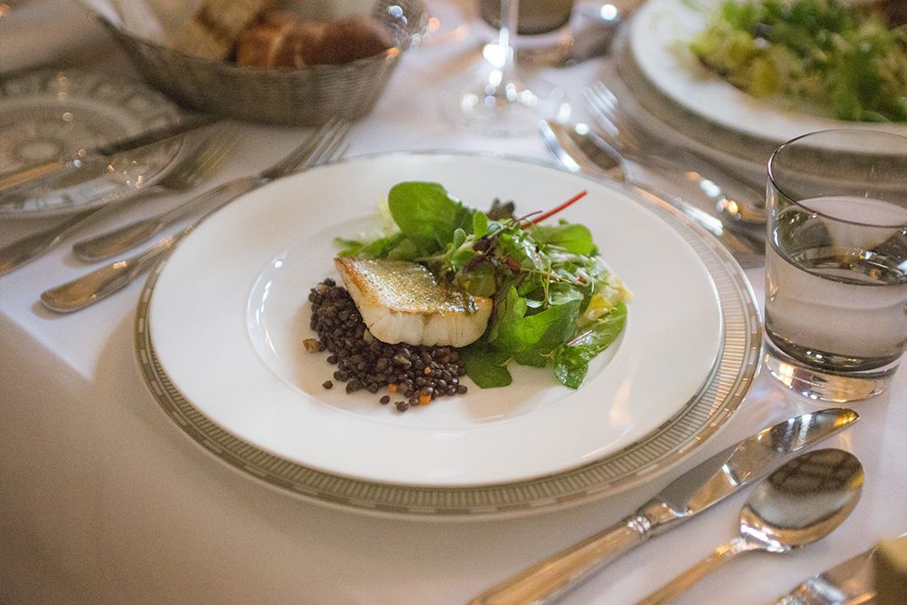 A plate with a piece of cooked fish, lentils and mixed leafy vegetables on a white tablecloth, surrounded by silver cutlery and glasses.