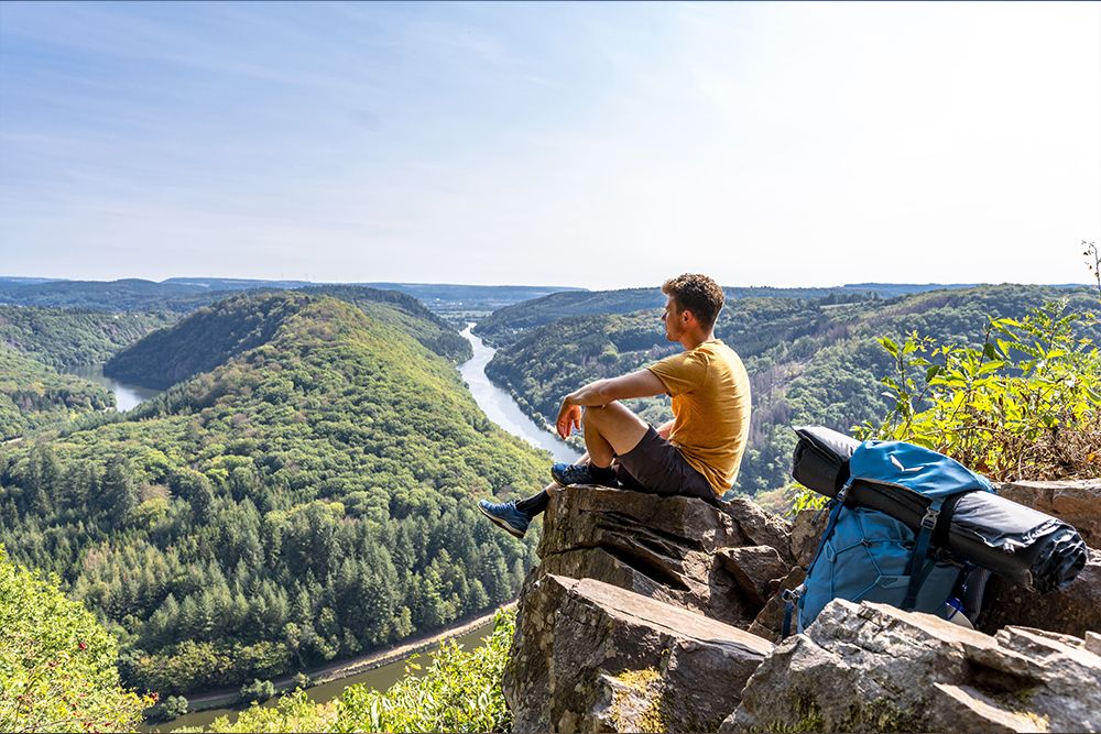 Eine Person sitzt in legerer Outdoor-Kleidung auf einem Felsvorsprung und blickt auf das malerische Saarlandtal mit Fluss und dichten Wäldern. Neben ihr ruht ein blauer Rucksack unter dem klaren Himmel – ein perfekter Platz entlang des Premiumwanderwegs Traumschleifen.