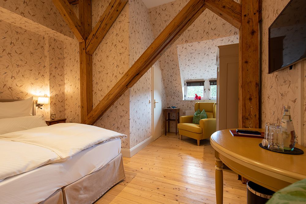 Gemütliches Schlafzimmer mit Holzbalken, Blumentapete und einem Bett. Kleiner Sitzbereich mit Sessel in der Ecke neben einem Fenster.