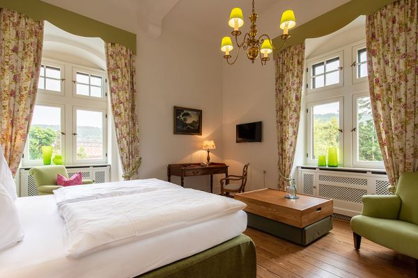 Das Hotelzimmer im Schloss Saareck verfügt über ein großes Bett, geblümte Vorhänge und einen eleganten Kronleuchter. Durch die Bogenfenster ist die Aussicht bezaubernd. Zu den zahlreichen Annehmlichkeiten gehören ein Holzschreibtisch, ein an der Wand montierter Fernseher und ein grüner Sessel.
