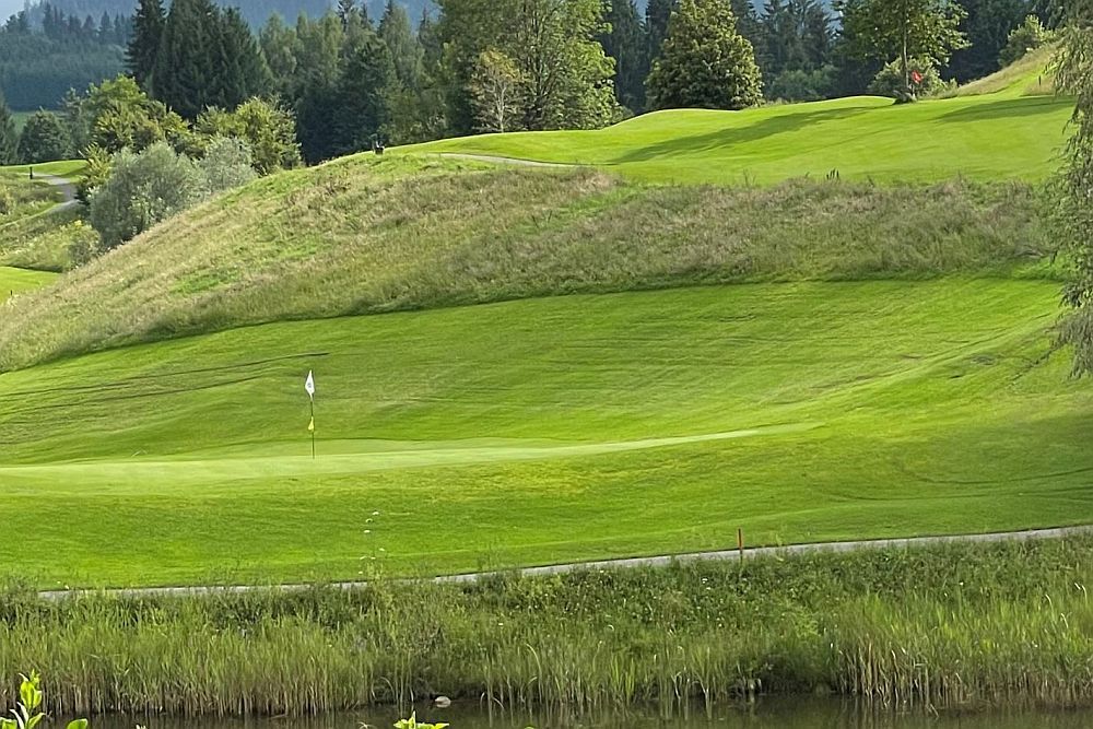 Ein üppiger Golfplatz bei Schloss Saareck bietet sanfte grüne Hügel, eine Fahne auf dem Grün und Bäume im Hintergrund. Ein kleiner Teich ziert den Vordergrund, perfekt für anspruchsvolle Golfsportler.