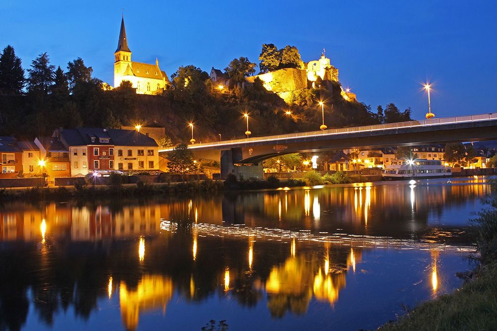 Church and illuminated bridge reflected in a calm river at night, with buildings and trees on a hill in the background.