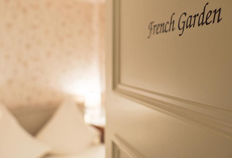 Die Tür mit der Aufschrift „Französischer Garten“ im Schloss Saareck steht leicht offen und lässt den gemütlichen Charme des Inneren erahnen. Sanftes, warmes Licht wirft einen sanften Schein über ein Bett und lädt die Gäste ein, in diesem eleganten Saar-Hotel Ruhe und Gelassenheit zu erleben.