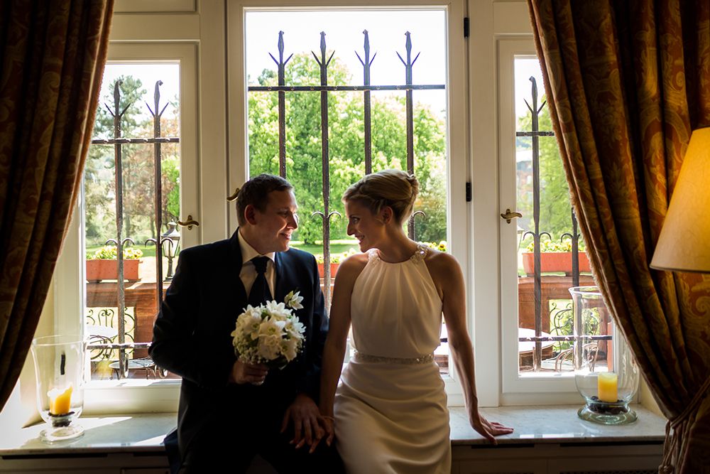 The bride and groom are sitting at a window with iron bars and holding flowers in their hands. They are facing each other and smiling, dressed in formal attire. The outside view shows green spaces and a balcony with planters.
