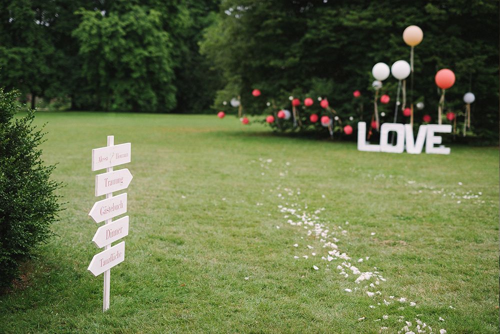 In einer märchenhaften Rasenlandschaft weist ein Wegweiser die Gäste zu verschiedenen zauberhaften Orten. Luftballons und große „LOVE“-Buchstaben schaffen eine traumhafte Kulisse, während weiße Blütenblätter zart auf dem Rasen verstreut sind – perfekt für eine Traumhochzeit.