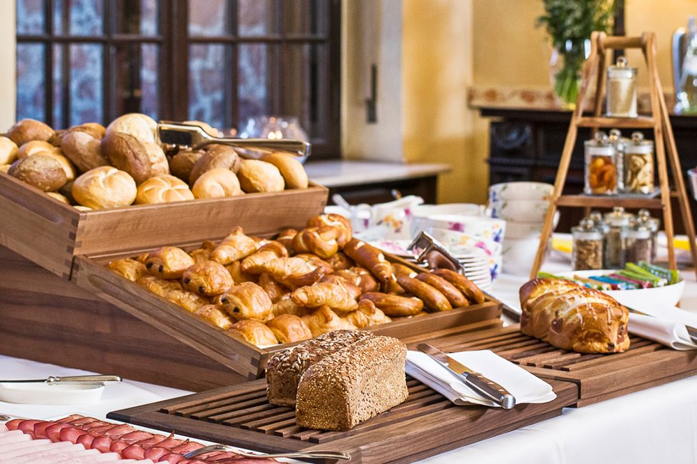Ein Frühstücksbuffet besteht aus einem Buffettisch mit verschiedenen Brotsorten, Brötchen, Gebäck und einem Laib Brot auf Holztabletts. Besteck und Utensilien begleiten das Buffet. Im Hintergrund sind Gläser und eine grüne Pflanze zu sehen. In der Nähe genießen Sie Kaffee oder Tee für den perfekten Start in den Tag.