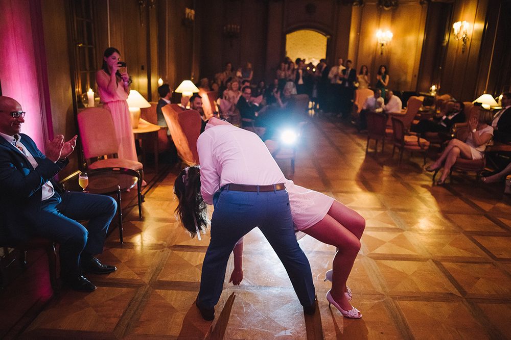 A couple performs a dramatic dance dip on a wooden floor in front of seated and standing guests in a warmly lit room.