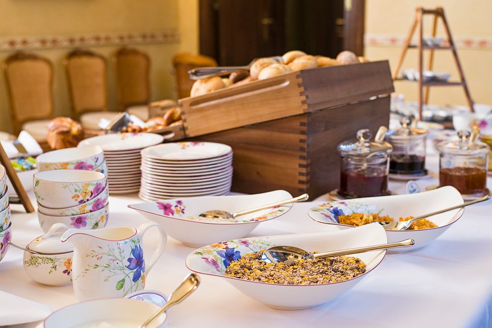 Ein Tisch ist mit Geschirr mit Blumenmuster, Schüsseln mit Müsli, Marmeladengläsern, Butter und einer Holzkiste mit verschiedenen Brötchen gedeckt – alle Köstlichkeiten für ein einladendes Frühstücksbuffet.