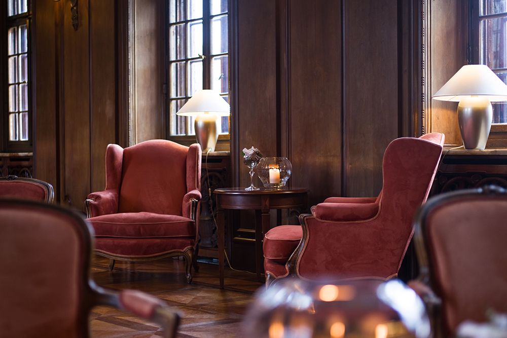 Ein schwach beleuchtetes Kaminzimmer verfügt über rote Samtsessel, einen kleinen runden Tisch mit einer Kerze und zwei Lampen an einer holzgetäfelten Wand. Es ist die perfekte Umgebung, um ein Glas Single Malt Whiskey zu genießen.