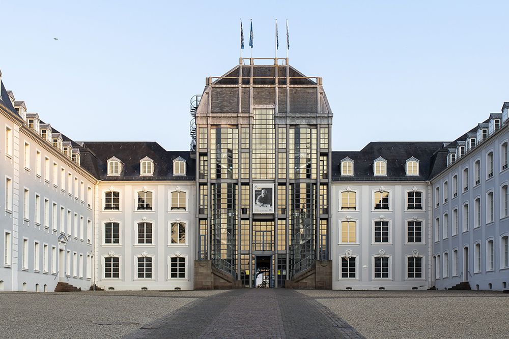 Symmetrical building with a glass centre section, surrounded by white wings, under a clear sky.